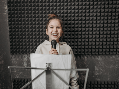Girl with her hair up and a white hoodie, holding a microphone with a music stand in front of her. 