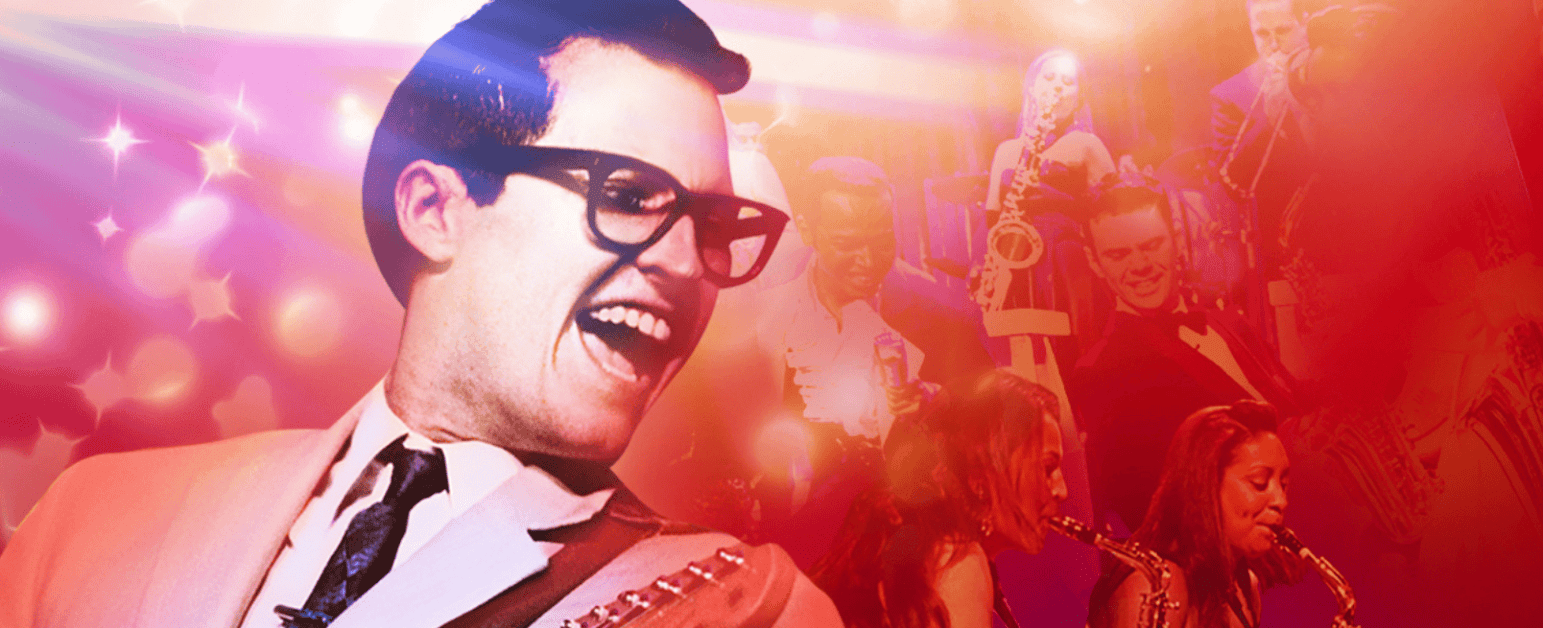 A performer dressed as Buddy Holly with short blakc hair, black rimmed glasses and a white suit holds a guitar in front of a background surrounded by performers