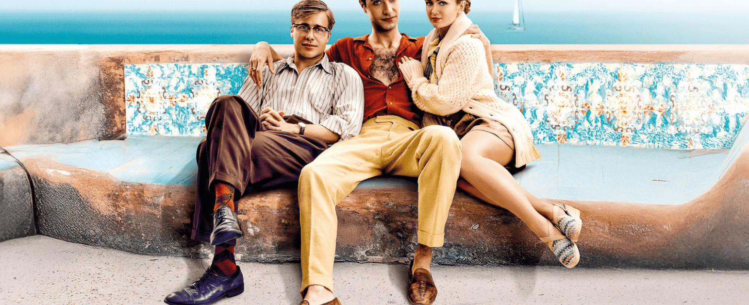 three people sitting on a bench with the sea in the background. The person in the middle has their arms around the two sat either side of them