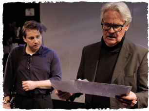 two actors wearing dark clothes looking at a piece of paper