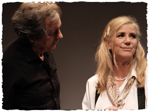 An actor with long blonde hair looking at the camera and an actor with short dark hair wearing a black t-shirt looking at them