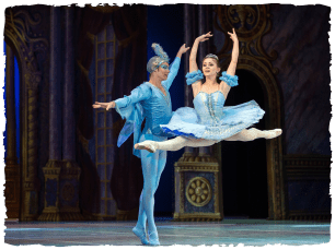 Two performers in blue ballet outfits, one jumping and raising their arms, the other standing behind them