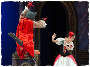 A performer dressed as a cat with a red cape jumps towards another performer in a black and white dress with a red bow