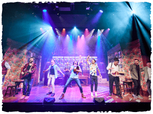 Bright lights. Group of men singing out to audience, playing instruments.