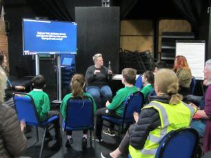 A group of school children sit around a screen where somebody is teaching them to sing in sign language.