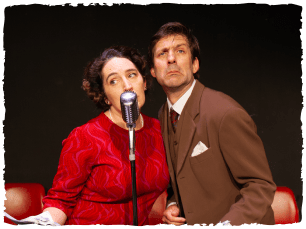 Woman in red dress and man in brown suit both looking out in to audience, talking in to same mic