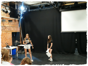 Two young people stood in a black box theatre space rehearsing a script