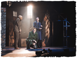 A man in a green trencoat kneeling and looking  mysterious while two men deliberate behind him