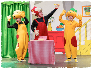 Actors on stage dressed as spot the dog and another dressed as a monkey in the middle standing behind a table 