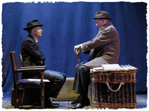 two actors sitting facing each other. One is sitting on a chair and one on a large wicker basket