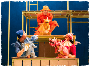 three actors dressed as dragons, holding dragon puppets (blue, pink and orange) all chatting to each other