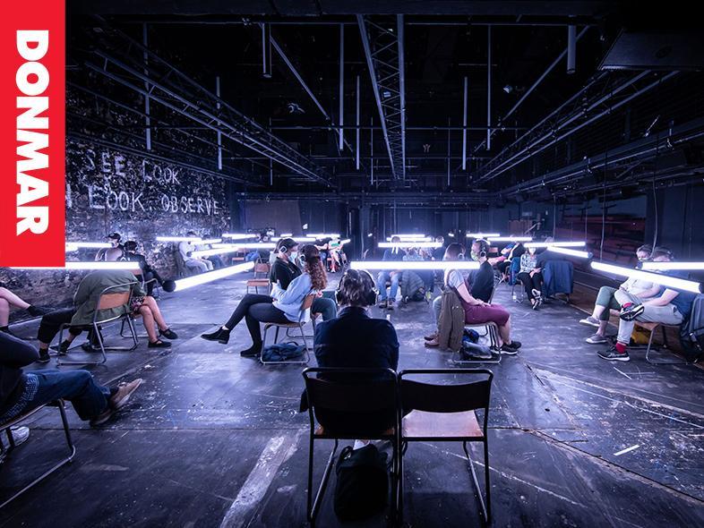 A group of audience members sit socially distanced in a darkened room, with halogen strip lights hovering in front of their faces. They all wear headphones and are sat in pairs, facing away from each other. It appears as if they are sat in a warehouse, with graffiti on the walls.