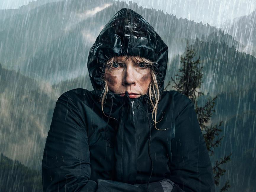 A blonde woman stands in a dark blue anorak, soaking wet, as it rains. She stands in front of a grey landscape of mountains and trees.
