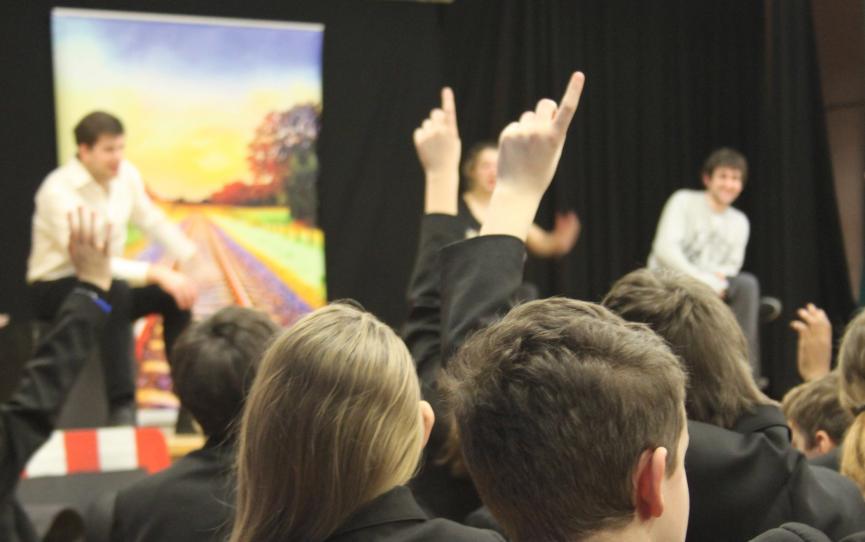 A group of school children sit listening to a talk. Some have their hands raised.