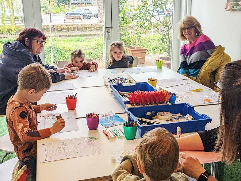 A group of adults and children sit around a large white table doing crafting activities. Out the window you can see a river and trees.