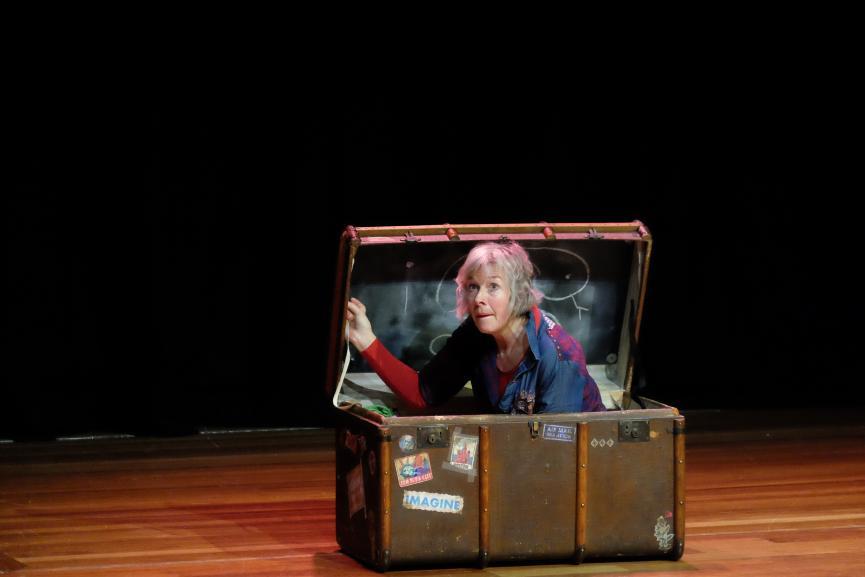 A woman sits in a chest on stage.