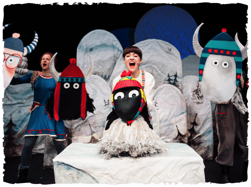 Performer in centre with a Yak puppet. Performers all singing. Cardboard Yak cut outs being held up around stage