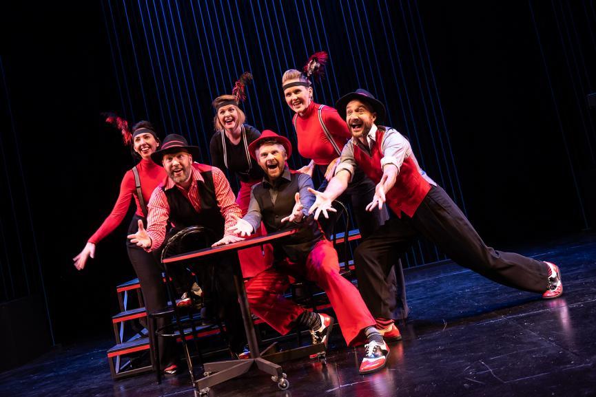 A cast of actors dressed in red and black stand on a dark stage all posing.