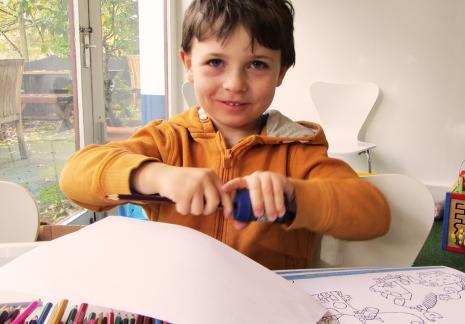 A young boy in an orange jumper sits at a table with a colouring book and crayons. He is sharpening a pencil with a blue pencil sharpener.