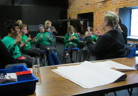 A group of school children in green uniforms sit around in a circle being taught how to use sign language in song.