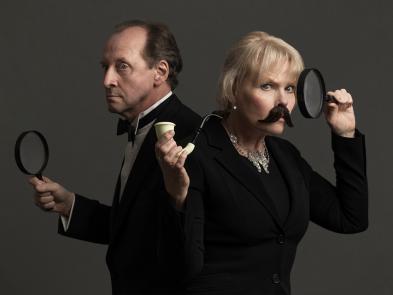 A man and a woman dressed in dark suits hold magnifying glasses and a pipe. The woman has a moutache.