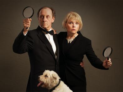 A man and a woman dressed in dark suits hold magnifying glasses and a pipe. A small white dog sits in front of them, completely unawares.