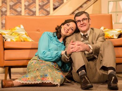 Joe Pasquale and Sarah Earnshaw sat cuddled together in front of an orange sofa in a room decorated in 1970s style.