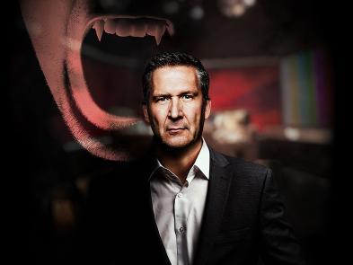 A man in a suit stands in a darkened television studio. There is a ghostly apparition appearing behind him with an open mouth and sharp teeth showing.