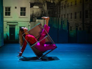 A woman in a pink top and trousers is trapped in a hollow metal-framed box which is tipping over backwards.