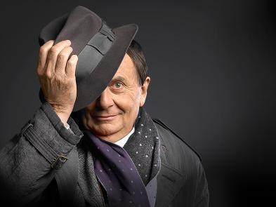 Barry Humphries in front of a black background, wearing a black suit and top hat. His hand is up lifting his hat slightly off his head.