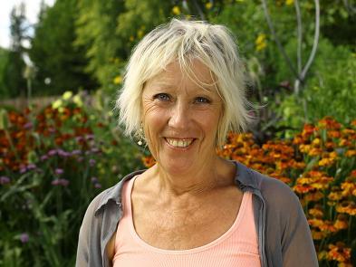 A woman with white hair, a pink top and a grey cardigan stands in a garden in front of green shrubs and dark red flowers.