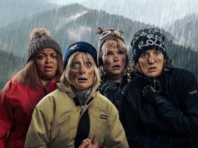 Four women in outdoor gear stood huddled together as they stand in a grey mountainous terrain as it rains.