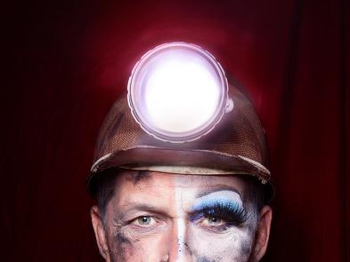 A miner wears a helmet with a large white light on the front. Half of his face is done up in make up.