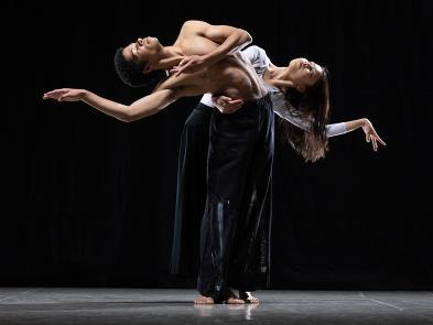 Two dancers stand in a black studio, they stand together but have arched backwards away from each other.