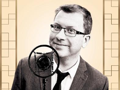 A man wearing a suit and glasses stands behind an old style radio mirophone