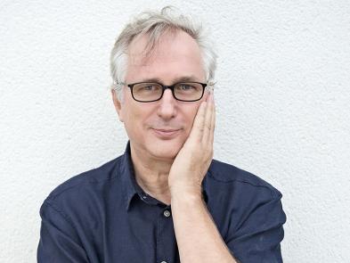 A man with grey hair and glasses stands in front of a plain background, holding his head in his hands.