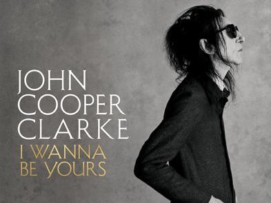 John Cooper Clarke dressed in black in front of a grey background.