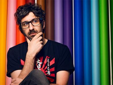 Mark Watson sits with his head in his hands in front of a rainbow background.