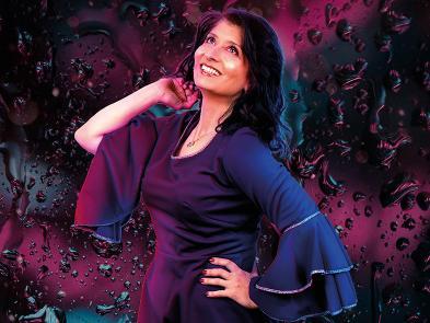 Shaparak Khorsandi stands in a purple dress, one hand on her hip, the other touching her next. She stands in front of swirling background of black, blue and purple.