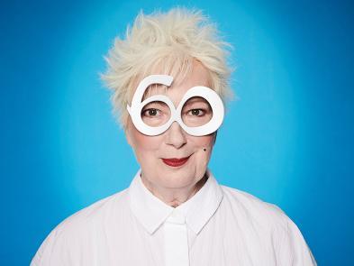 Jenny Eclair, a woman with short white hair stands in front of a blue screen with a white buttoned up shirt on. She is wearing a pair of white glasses, where the lenses are in the shape of the number 60.