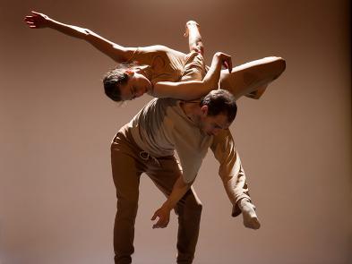 Two dancers mid-pose. One lies across the back of the other, who is hunched over.