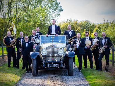 A group of performers and musicians in tuxedos stand around a vintage car on a country lane