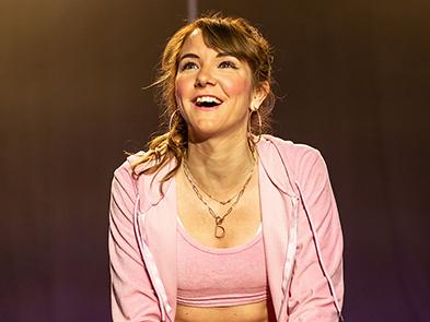A young white woman with brown hair in a jaunty high ponytail sits on a dark stage, lit by fluorescent pink LED lights. She wears a pink tracksuit, white Nike trainers, pink nail varnish and a gold 'D' necklace. She wears bright pink blush, cat eye eyeliner and thickly pencilled eyebrows. 