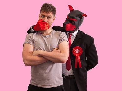 A man stands in a beige t-shirt in front of a pink background. His mouth is covered by a man dressed in a leather kink dog outfit and a suit with a red rosette.