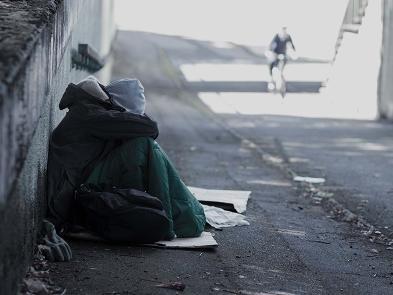 A person sits in a coat and sleeping bag with their head in the hands on an empty street.
