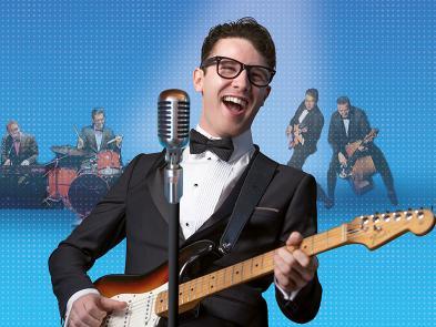 A man in a black and white suit with a bow tie plays the guitar. He is singing and wearing glasses. Behind him are various band members playing instruments.
