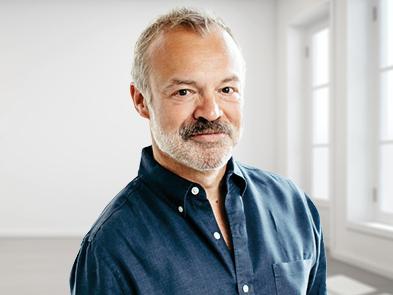 Graham Norton stands in a light room next to large windows, wearing a dark blue shirt.