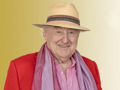 Henry Blofeld wears a red jacket and pink scarf with a straw trilby hat in front of a gold background.