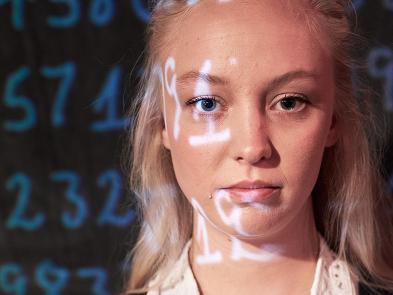 A young blonde girl looks forward in front of a black background. There is a projection of a series of numbers over the background and half her face.