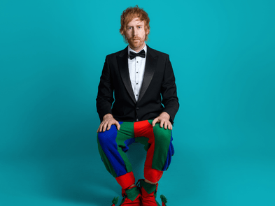 Mark Simmons sits in front of a blue backdrop in a black and white suit jacket but with colourful red blue and green medieval jester leggins and shoes